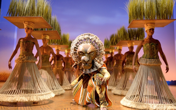 Lion King stage performance with actors in traditional costumes and lion mask.