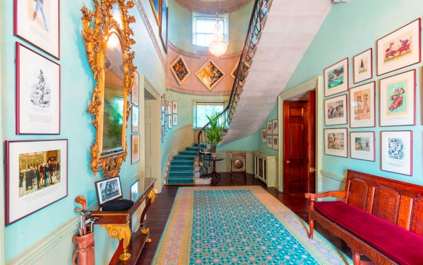 Hillsborough Castle interior with ornate mirror, framed art, and curved staircase.