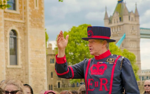 Beefeater guiding tourists at the Tower of London with Tower Bridge in the background.