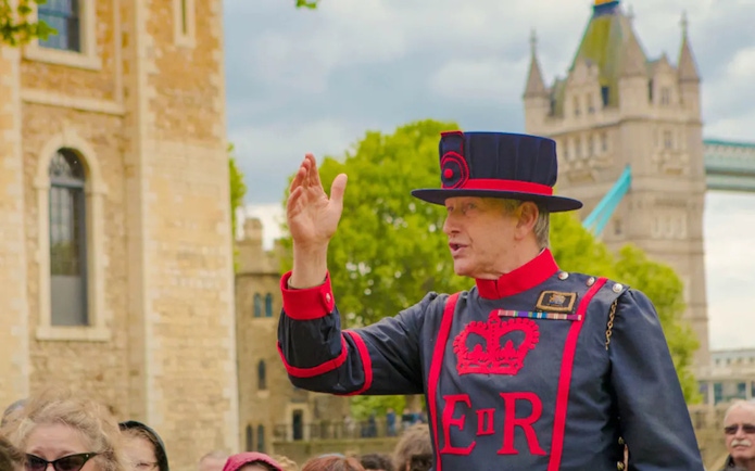 Beefeater guiding tourists at the Tower of London with Tower Bridge in the background.