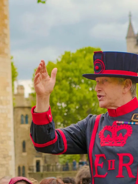 Beefeater guiding tourists at the Tower of London with Tower Bridge in the background.