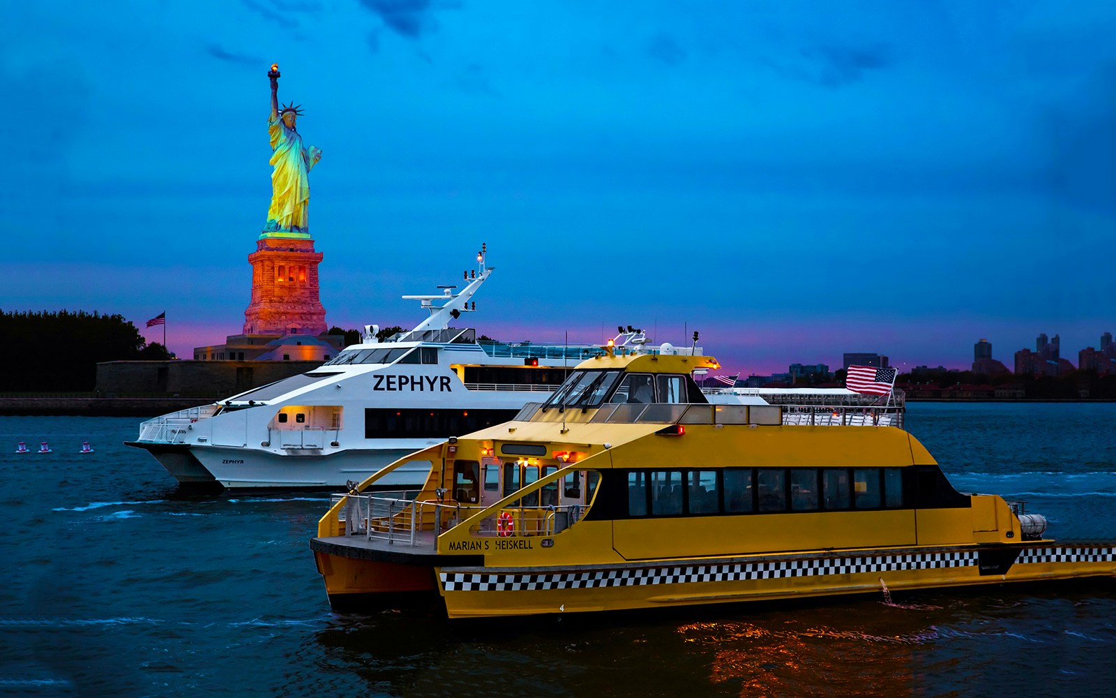Statue of Liberty illuminated at night with cruise boats in New York Harbor.