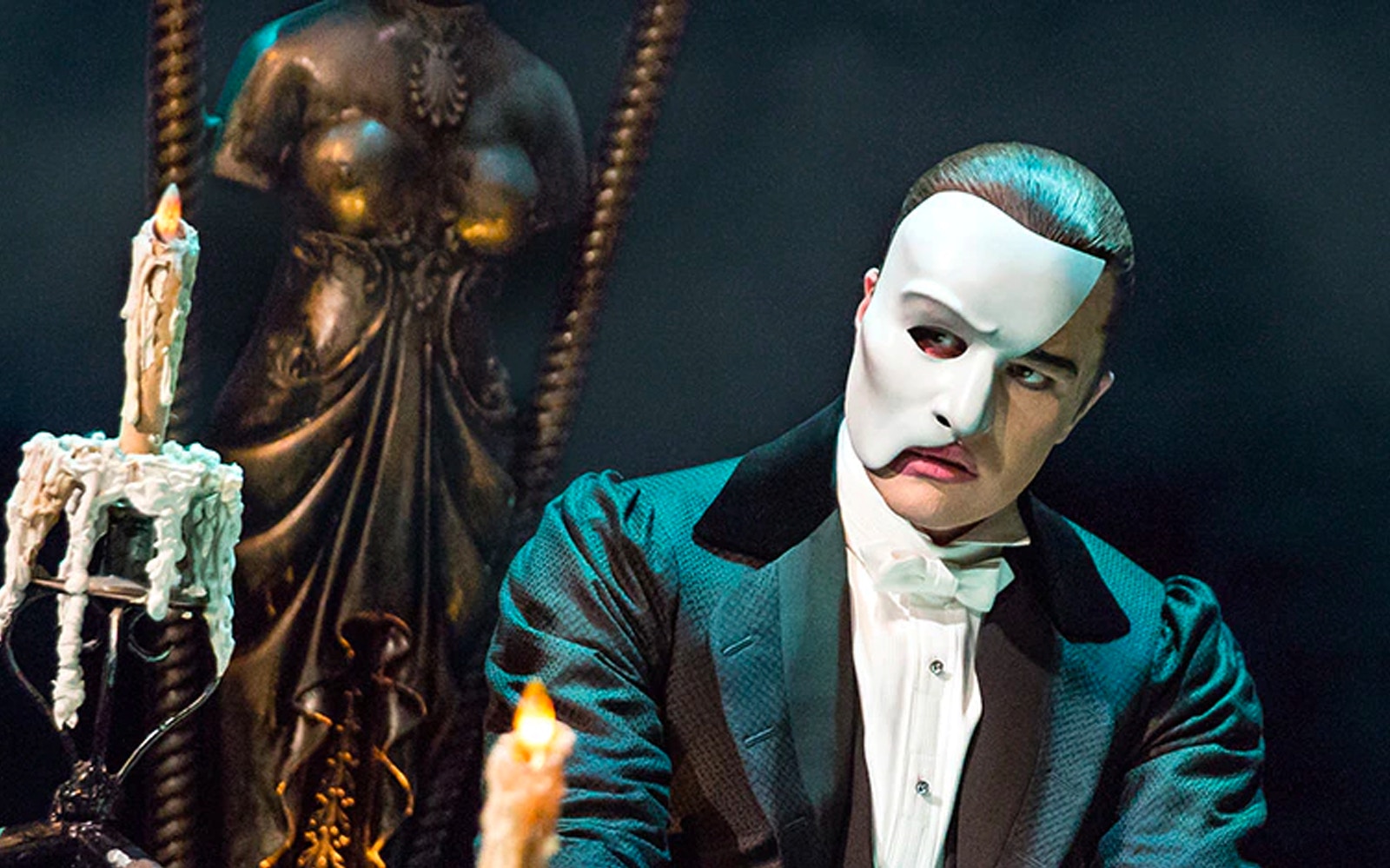 Phantom of the Opera character in mask with candles and statue in background.