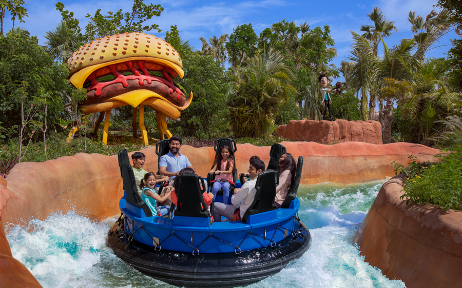 Group enjoying water ride at MOTIONGATE™ Dubai with giant burger sculpture in background.