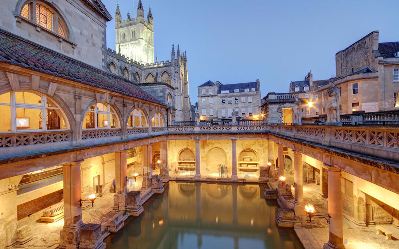 Roman Baths in Bath, England, illuminated at dusk with Bath Abbey in the background.