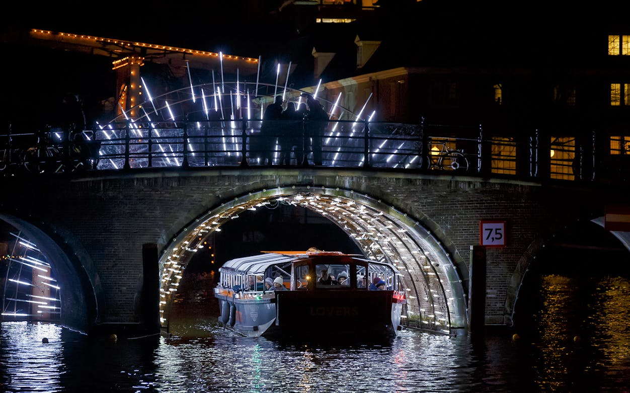 Cruise boat under illuminated Amsterdam bridge at night.