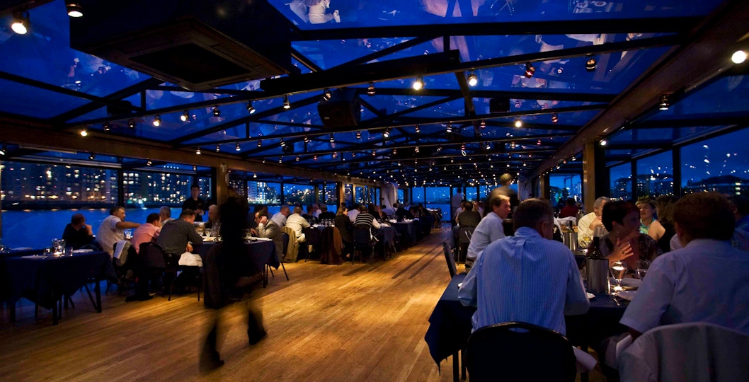 Guests dining on a Thames riverboat with city lights visible through large windows.