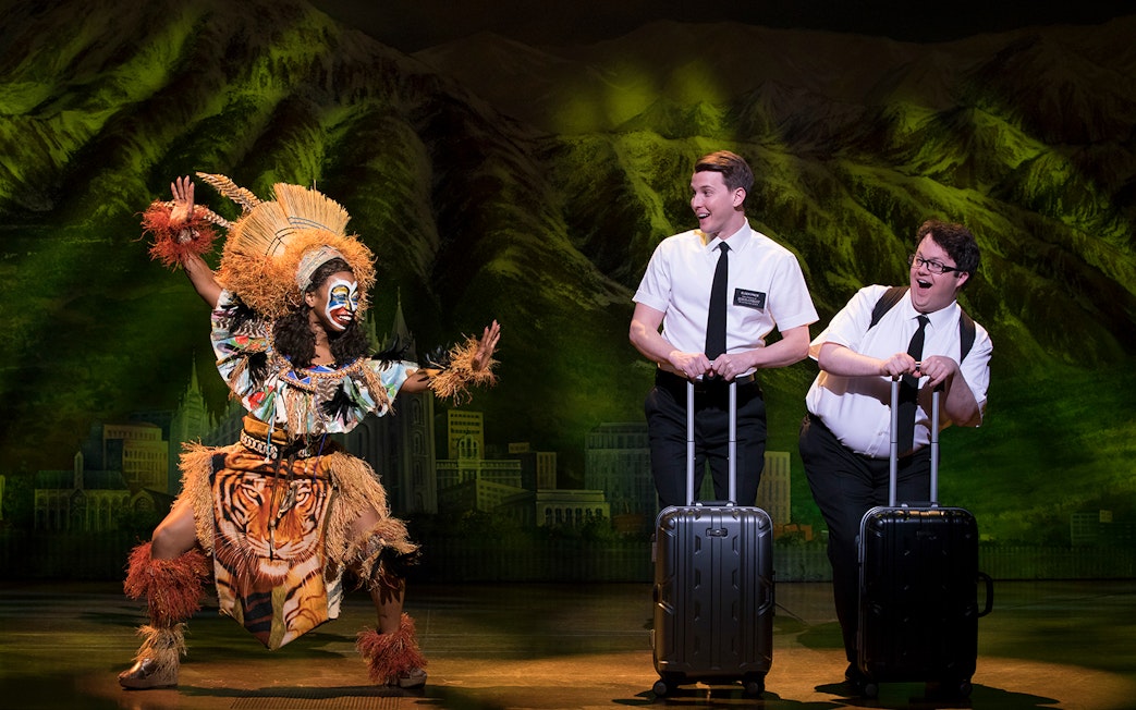 Performers in "The Book of Mormon" musical scene with vibrant costumes and props.
