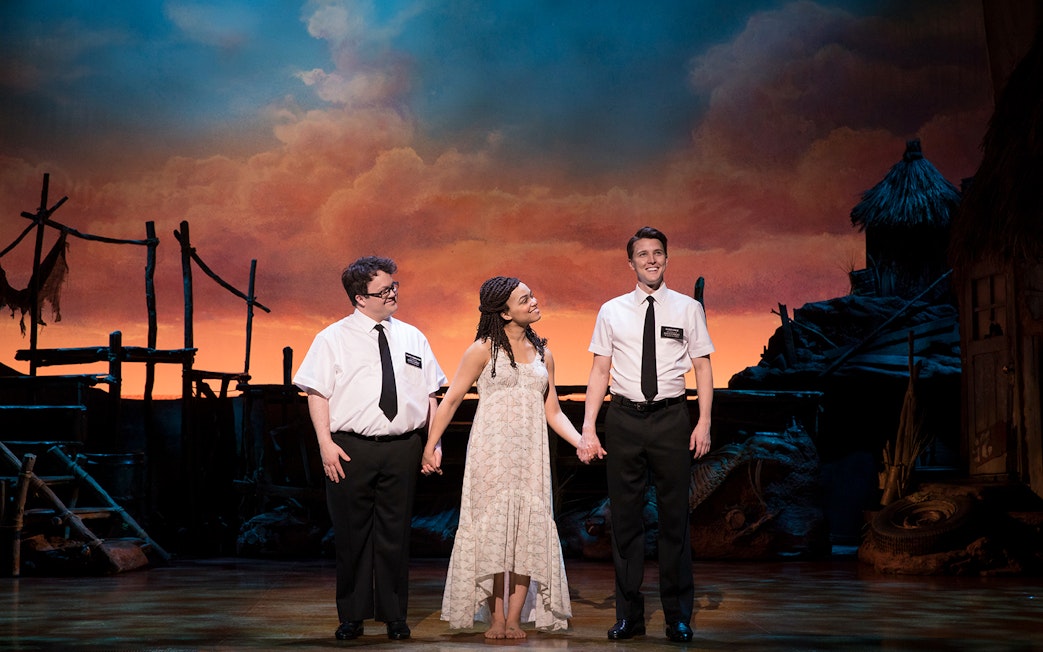 Performers on stage during "The Book of Mormon" musical with a sunset backdrop.