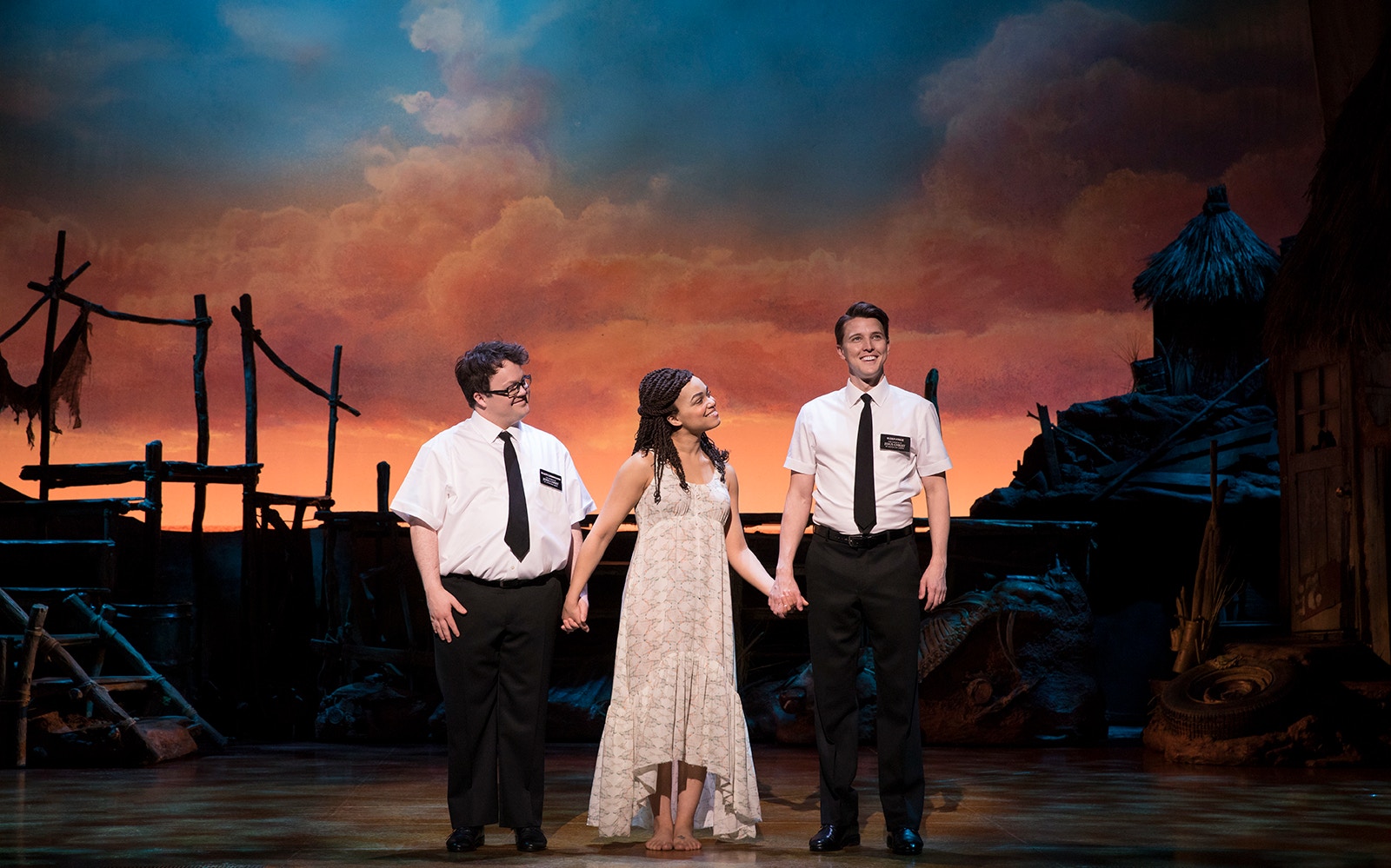 Performers on stage during "The Book of Mormon" musical with a sunset backdrop.