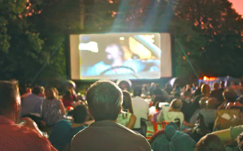 Crowd watching a movie at an outdoor cinema in a park at dusk.