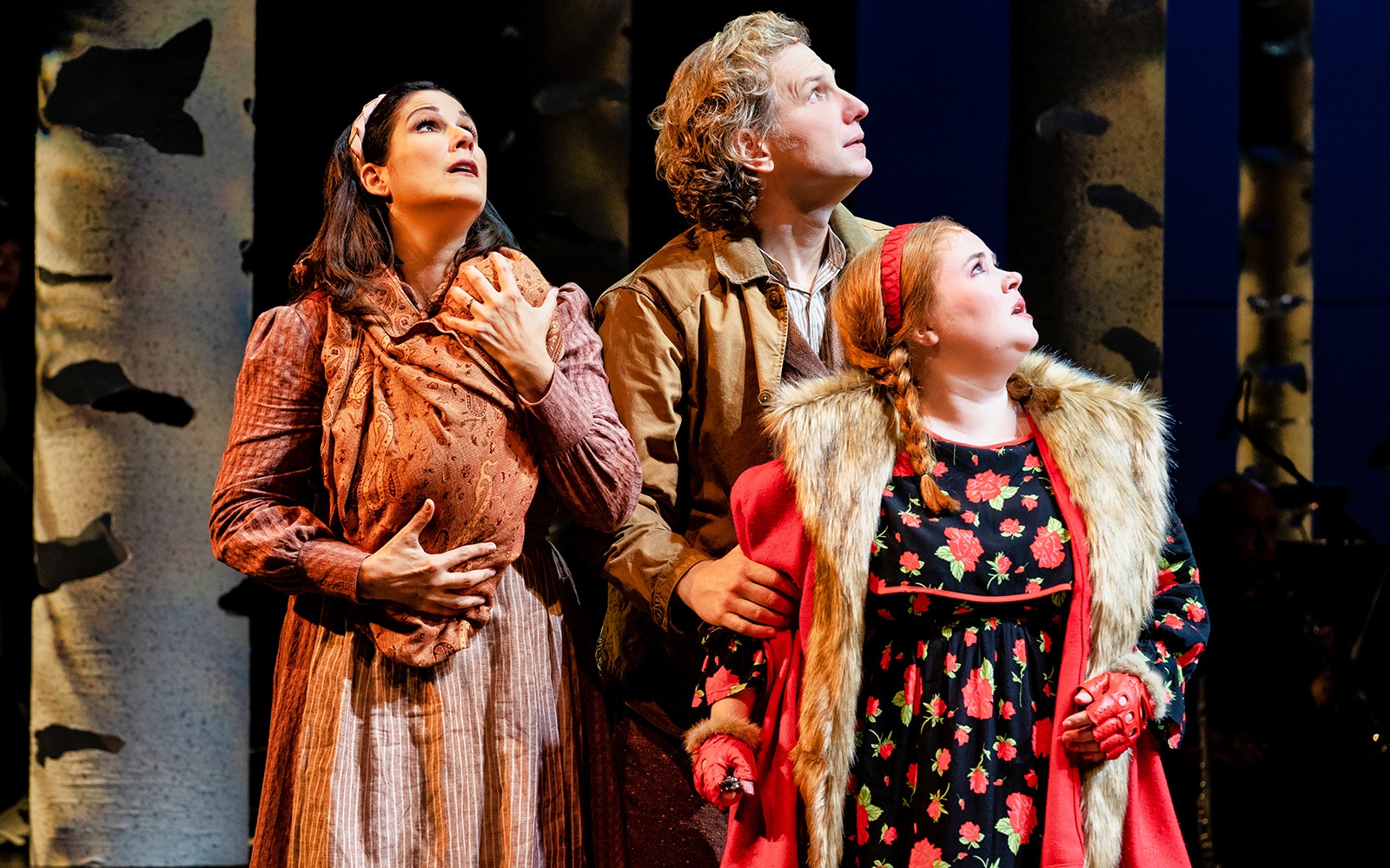 Three actors in period costumes look upward in a forest setting, part of "Into The Woods" performance.