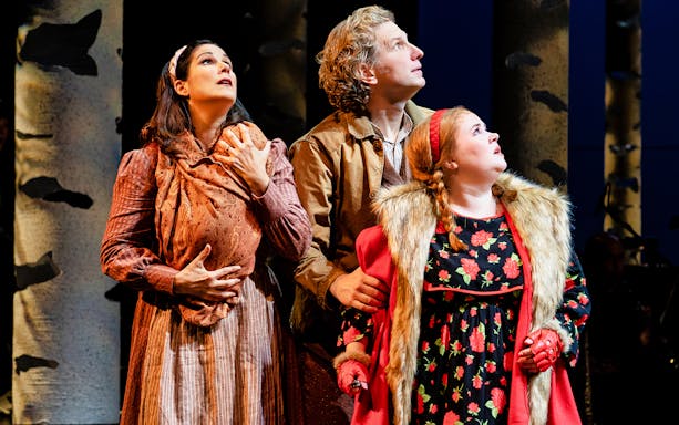 Three actors in period costumes look upward in a forest setting, part of "Into The Woods" performance.